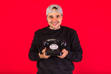 Man In A Cap Dressed In Black, Holds A Retro Telephone From The 60s And Smiles, On A Red Background, With Copy Space. Concept Of Contact, Social Relations And Phone Calls