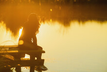 Woman Standing At The Edge Of A Deck And Starring At The Sun During A Beautiful Sunset By The Lake. Photo As Concept For Serenity And Meditation.