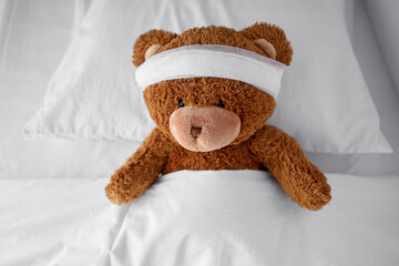 Wall Mural - medicine, healthcare and childhood concept - ill teddy bear toy with bandaged head lying in bed
