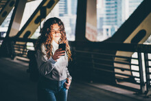 Curly Haired Young Woman Using Phone With Backpack Walks Under Bridge.