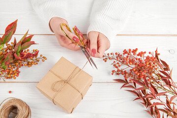 Wall Mural - Top view of female hands wrapping autumn creative holiday present. Handmade paper gift box with foliage leaves. Thanksgiving day, fall background. top view, flat lay. Holiday and sales season