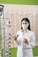 Wall Mural - asian oculist in medical mask comparing eyeglasses while working in optics shop.