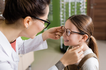 Sticker - oculist putting eyeglasses on cheerful girl in optics salon.