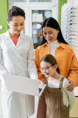 Poster - girl in eyeglasses looking at laptop near asian mom and smiling oculist in optics store.