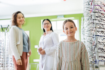 Poster - cheerful girl looking at camera near blurred mom and asian ophthalmologist in optics store.