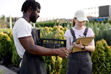 Black And Caucasian Farmers Talking, Discussing, Woman In Outdoor Greenhouse Taking Notes On Notebook, Examining Quality Of Plants Growing. In Modern Greenhouse, Working Together, Have Conversation