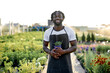 © Roman - Portrait of handsome black american gardener in uniform with green plants. Worker taking care of plants in outdoor garden, young positive bearded guy engaged in agriculture, flowering, gardening.