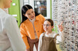 © LIGHTFIELD STUDIOS - positive kid choosing eyeglasses near asian mom and blurred ophthalmologist in optics shop.