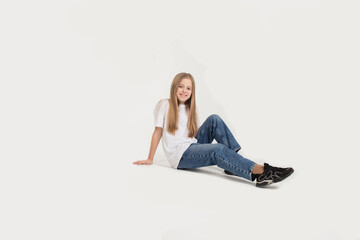 a beautiful teenage girl in a white T-shirt and blue jeans is sitting on a white background