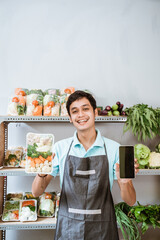 Wall Mural - Asian male greengrocer showing vegetables and showing a phone screen