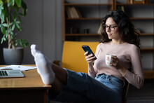 Smiling Millennial Female Employee Procrastinate On Workplace Sit With Cellphone And Feet On Desk. Girl Distracted From Computer Job Read Text Message On Smartphone Device Browsing Wireless Internet