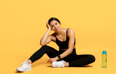 Wall Mural - Wellness and training. Happy fit young black lady sitting on the floor with bottle of water at yellow background
