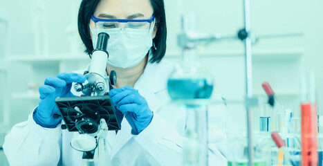 Wall Mural - Studio shot of Asian professional female middle aged scientist in white lab coat safety glasses face mask and rubber gloves using microscope lens looking at covid19 coronavirus sample in laboratory