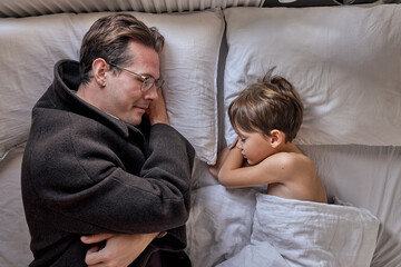 Wall Mural - Father come to say goodbye to child boy before leave for work in morning, on bed. top view on sleepy man and kid at home, adult male in coat, in eyeglasses. family, monday, lifestyle concept