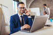 © Mediteraneo - Happy businessman using laptop and phone with team discussing project in the background.