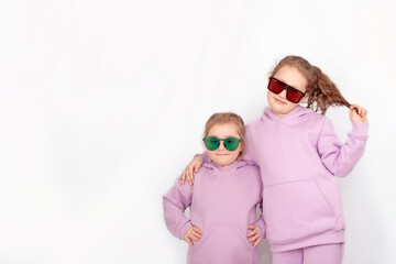 Two little beautiful smiling girls in fashionable spring clothes with a hood and sunglasses.  Carefree children in identical hoodies pose on a white background.