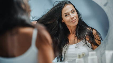 Wall Mural - Beautiful Early Middle Aged Woman Looks into Bathroom Mirror Touches Her Lush Black Hair, Admires Her Looks. Concept for Happiness, Wellbeing, Natural Beauty, Organic Skin Care Products