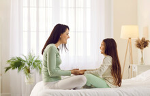 Happy Family Having Fun In Cozy Warm Bedroom. Cheerful Young Mother And Child In Comfy Homewear Sitting Together On Bed At Home, Looking At Each Other, Holding Hands And Smiling. Care And Love Concept