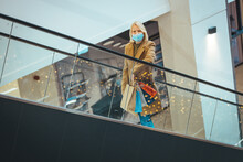 Young Stylish Woman With Protective Mask Climbs Escalator With Shopping Bags In Shopping Center. Entertainment Infrastructure In Shopping Centers Concept
