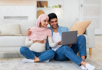 Wall Mural - Loving pregnant arab spouses using laptop, sitting on floor at living room, watching baby room interior, free space