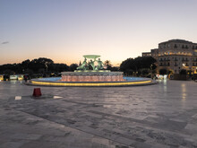 The Triton Fountain Lit Up In Triton Square With The Phoenicia Malta Hotel And Sun Setting In The Background, Valletta, Malta.