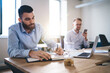 © BullRun - Caucasian man in formal t shirt writing strategy ideas in textbook sitting at table desktop with male colleague using smartphone gadget for phoning on blurred background, office working process