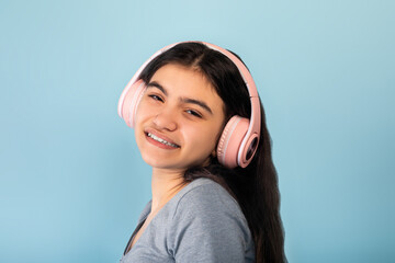 Wall Mural - Indian teen girl in casual wear listening to music in pink wireless headphones, enjoying cool song on blue background