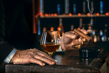 Man's Hands With A Cigar, Elegant Glass Of Brandy On The Bar Counter. Alcoholic Drinks, Cognac, Whiskey, Port, Brandy, Rum, Scotch, Bourbon. Vintage Wooden Table In A Pub At Night