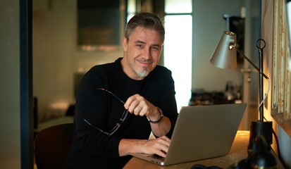 Older man sitting at desk in dark cosy room working on laptop computer in home office. Mature age, middle age, mid adult casual man in 50s, confident happy smiling.