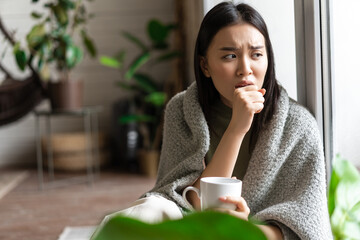 Wall Mural - Sad ill asian girl staying on self quarantine during covid, catching flu and sitting at home with cup of tea near window, coughing in hand