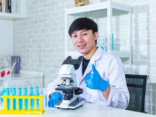 Sticker - Portrait studio shot of Asian young professional male scientist in white lab coat sitting smiling look at camera behind working laboratory desk full of test tubes beakers flasks sample and microscope