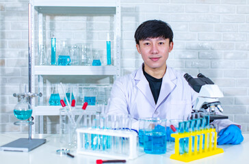 Sticker - Portrait studio shot of Asian young professional male scientist in white lab coat sitting smiling look at camera behind working laboratory desk full of test tubes beakers flasks sample and microscope