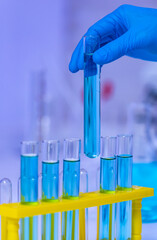 Wall Mural - Closeup studio shot of scientist hand wearing rubber gloves picking up blue reagent solvent solution liquid sample in glass test tube from yellow plastic rack in laboratory with effect light flare