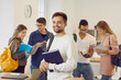 © Studio Romantic - Portrait of cheerful male university student in classroom. Handsome young man with happy face expression holding textbook and smiling at camera while his classmates are reading books in background