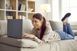 © Studio Romantic - Happy woman using her laptop computer. Smiling young girl lying on a comfy sofa at home, using her modern notebook PC, working remotely or studying a new online course