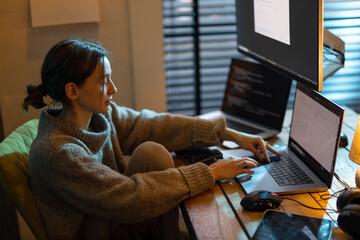 Young focused woman feeling overworked and tired, working on laptop and desktop computers, sitting at home office during the nighttime. Female programmer writing code