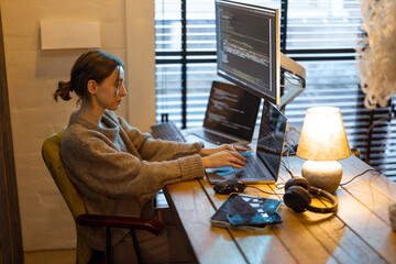 Young woman works on computers, sitting at workplace at cozy home office interior. Concept of freelance and remote work from home. Programmer writing code. Caucasian woman wearing domestic clothes.
