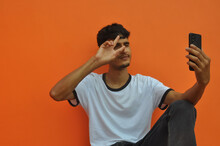 A Young Guy Showing Peace Sign While Taking Selfie, Sitting Against Orange Wall Background With Copy Space
