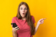 © luismolinero - Young woman using mobile phone isolated on yellow background surprised and pointing side