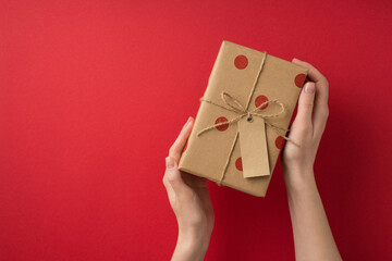 Wall Mural - First person top view photo of valentine's day decorations girl's hands demonstrating craft paper gift box with polka dot pattern pricetag and twine bow on isolated red background with copyspace