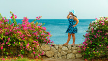 A Brunette Woman In A Blue Sundress And Hat Among Blooming Bougainvillea Stands On The Wall Against The Background Of The Sea.