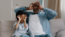 Happy Cheerful African American Mature Father Playing With Cute Daughter Sitting In Room On Sofa Smiling Dad And Little Girl Making With Hands Funny Glasses Masks Pretend Wearing Spectacles Fooling