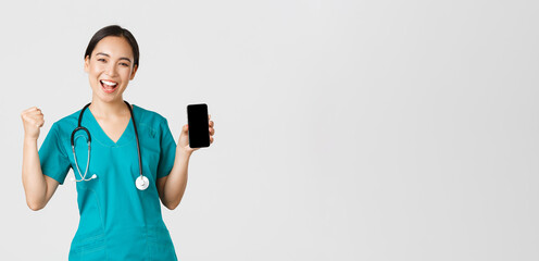 Wall Mural - Covid-19, healthcare workers and online medicine concept. Successful cheerful asian female doctor, nurse in scrubs fist pump in rejoice, showing smartphone screen app and looking pleased at camera