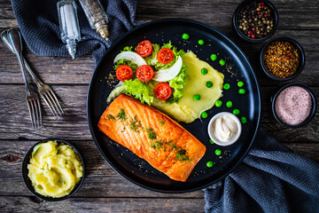 Fried salmon steak with potato puree and vegetable salad served on black plate on wooden table
