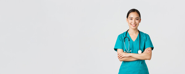 Wall Mural - Covid-19, healthcare workers, pandemic concept. Portrait of confident smiling, attractive asian female nurse in scrubs, with stethoscope, cross arms chest and looking at camera, white background