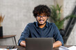 © Vadim Pastuh - Front view of positive Hindi man in smart casual shirt using laptop while sitting at the desk in his flat. Young Indian male student watching webinars, educational courses, learning on the distance