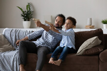 Excited Afro American Dad And Son Kid Playing Video Game, Using Toy Guns, Aiming Pistols Away, Sitting, Resting On Home Couch, Competing In Online Battle, Engaged In Virtual Shooting