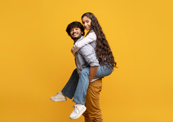 Wall Mural - Young indian man giving piggyback ride to his girlfriend against yellow studio background, free space