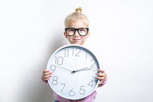 Smiling Child Girl In Glasses Holds A Large Wall Clock On A Light Background.