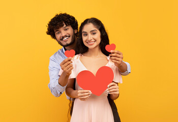 Wall Mural - Love is in the air. Romantic indian couple with red paper hearts in hands posing over yellow studio background